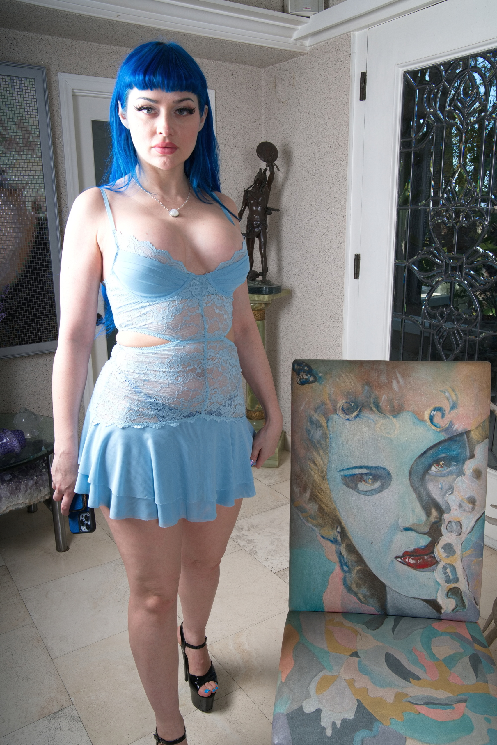 Woman with bright blue hair in a light blue lace dress stands indoors near a colorful painted chair with a stylized face.